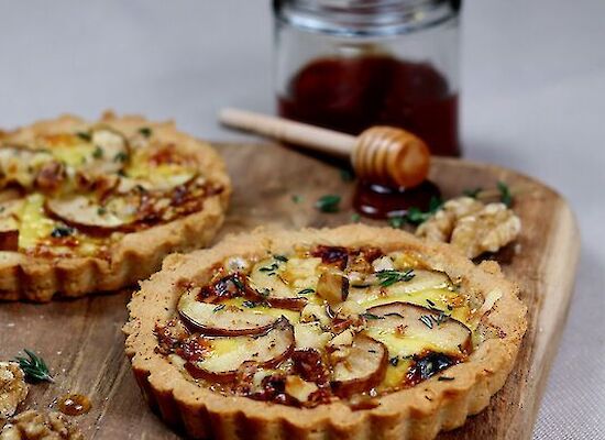 Tartelette mit Bockshornklee-Käse auf einem Holzbrett mit einem Honigglas und Honiglöffel.