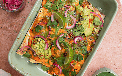Baking dish with chicken enchiladas, avocado, pickled red onion and melted jalapeño cow’s cheese, garnished with coriander and lime