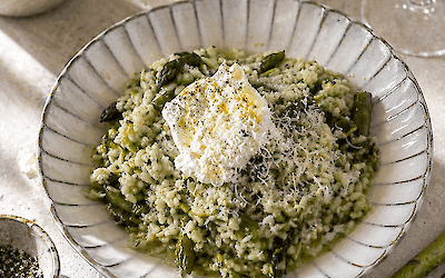 Creamy risotto verde with green asparagus, basil, and lemon, served with burrata and grated cheese on a light plate, with fresh asparagus and olive oil in the background