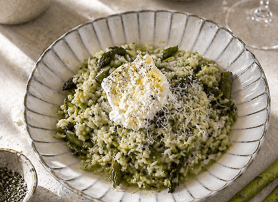 Cremiges Risotto verde mit grünem Spargel, Basilikum und Zitrone, serviert mit Burrata und geriebenem Käse auf einem hellen Teller, mit frischem Spargel und Olivenöl im Hintergrund