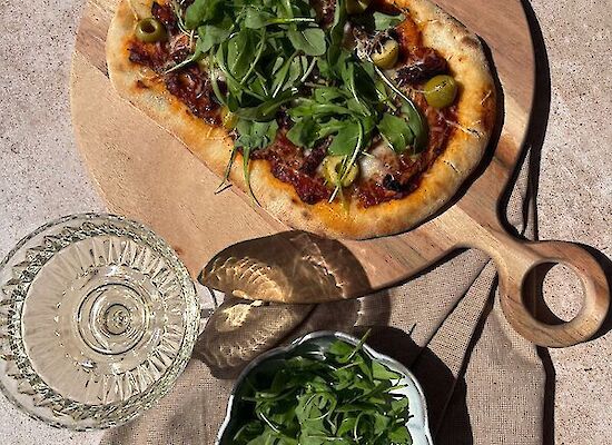 Pinsa au fromage de chèvre, roquette, olives et tomates séchées sur une planche en bois, accompagnée d’un verre de vin blanc et d’un bol de roquette